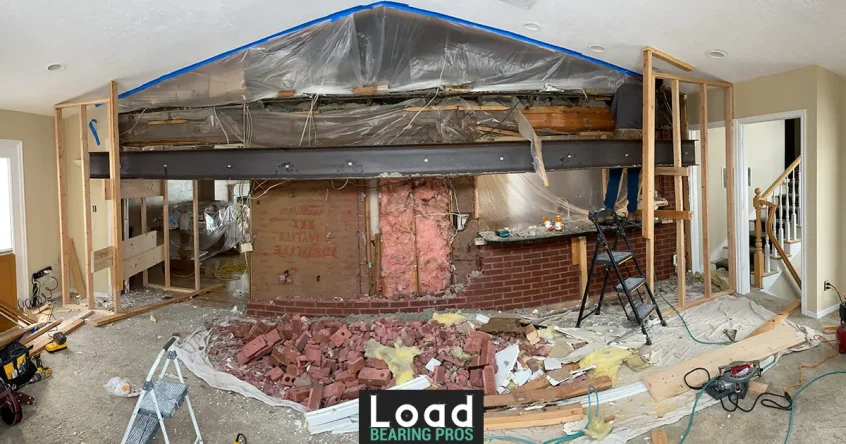 A black steel support beam installed mid-wall during a load-bearing wall removal, with exposed brick and torn-out drywall below.