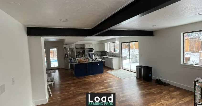 An open-concept kitchen and living area with a black steel support beam running across the ceiling after a load-bearing wall removal.