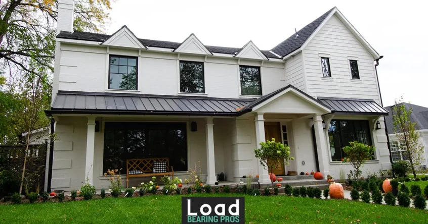 A white two-story farmhouse-style home with black standing-seam metal roof accents and pumpkin-lined landscaping.