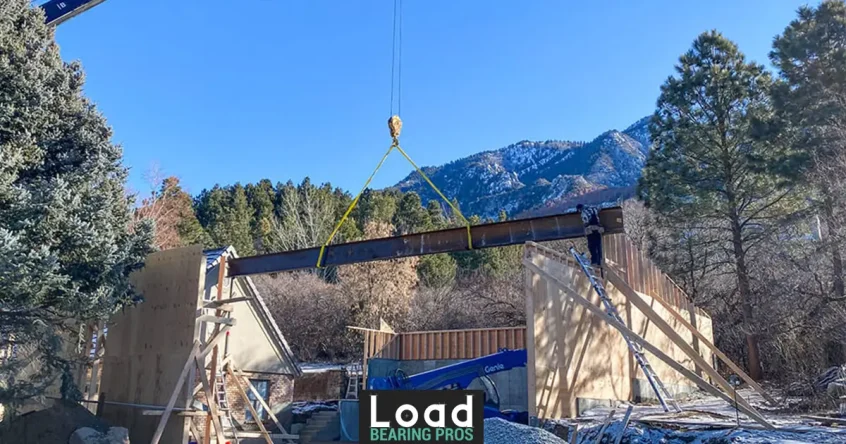 A crane lifts a large steel support beam into place above a home’s framed walls during construction, with mountains in the background.